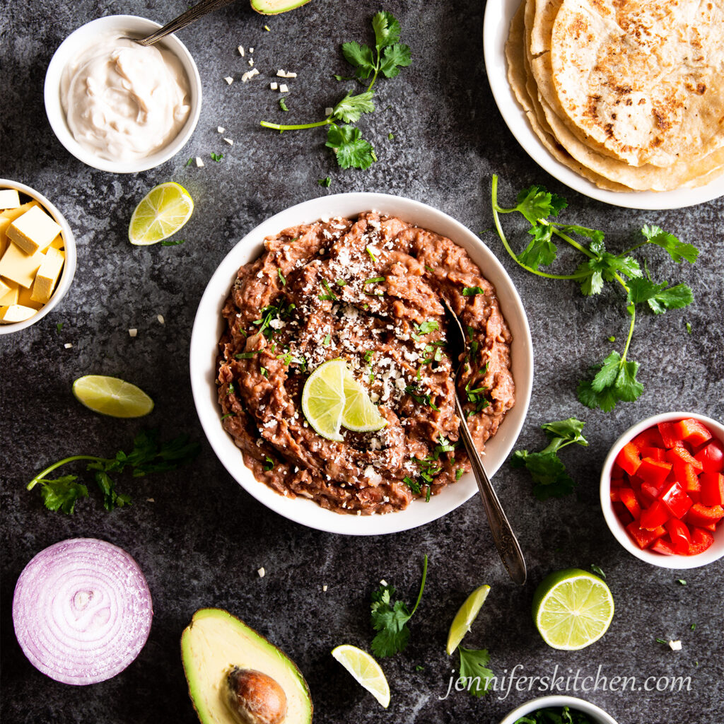 A bowl of refried beans with cilantro, lime wedges, avocado, peppers, red onion, and sour cream.