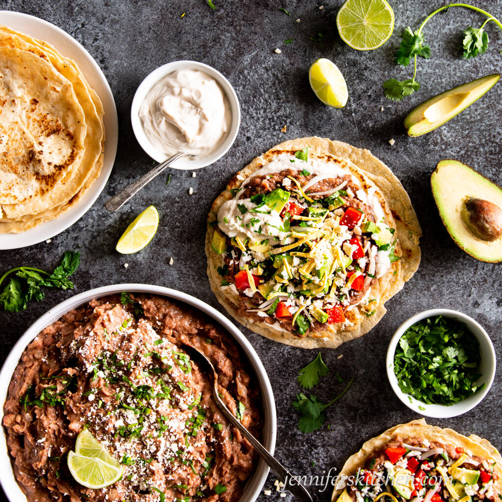 Tortillas topped with refried beans, cilantro, lime wedges, avocado, peppers, red onion, and sour cream.