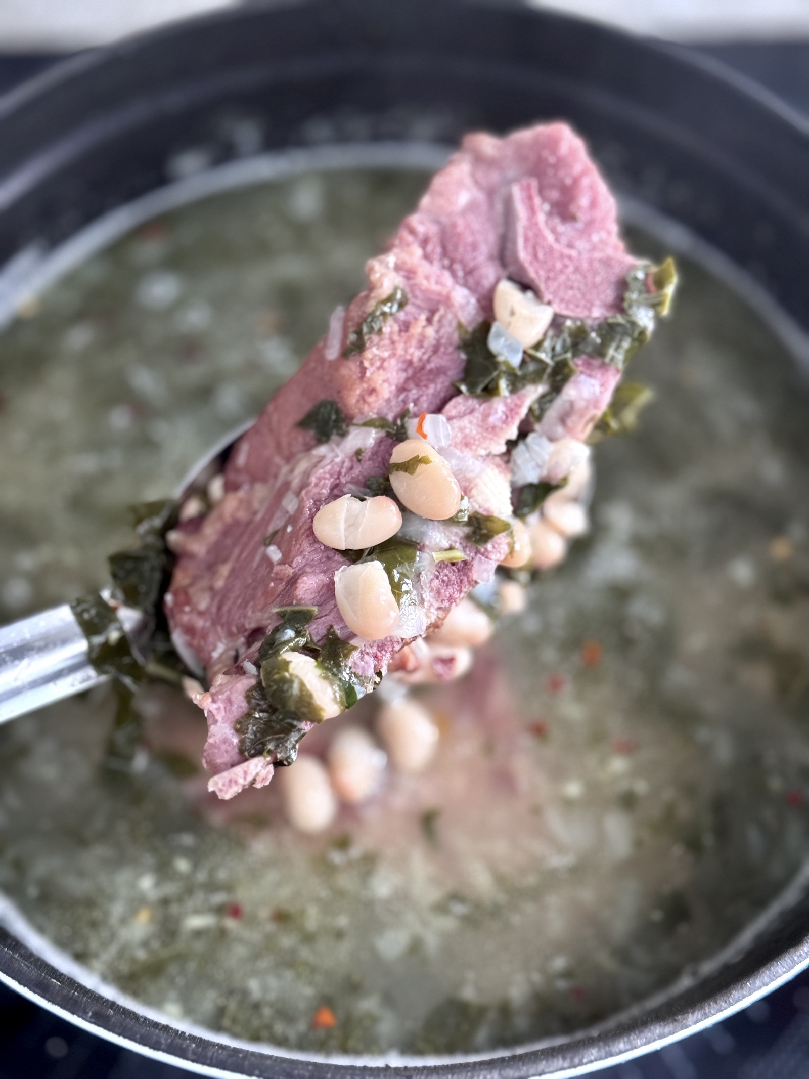 A pork neck bone on a spoon above a pot of bean soup