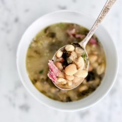 Top view of a spoonful of smoky white bean soup with ham and the white bowl on a marble background