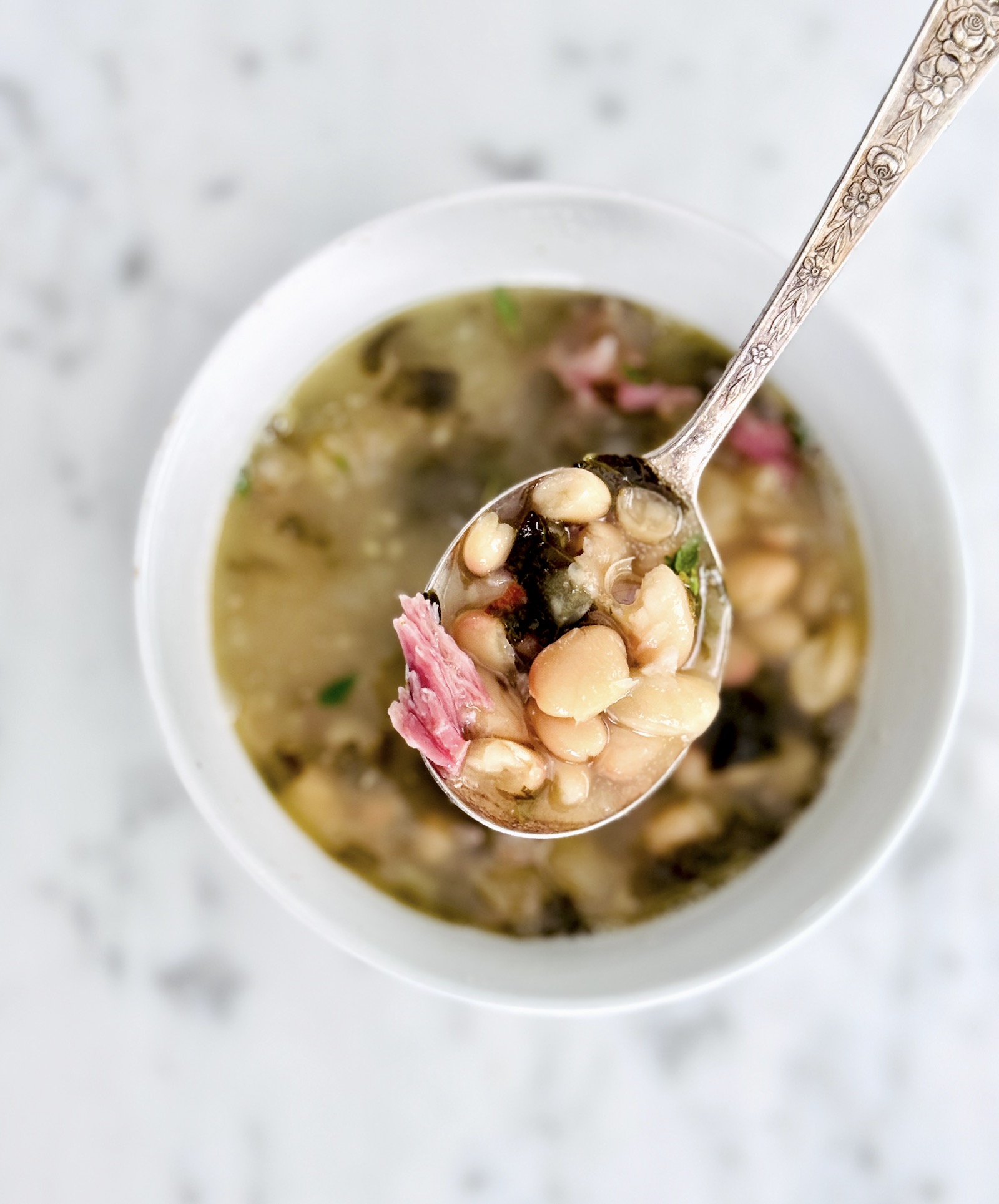 Top view of a spoonful of smoky white bean soup with ham and the white bowl on a marble background