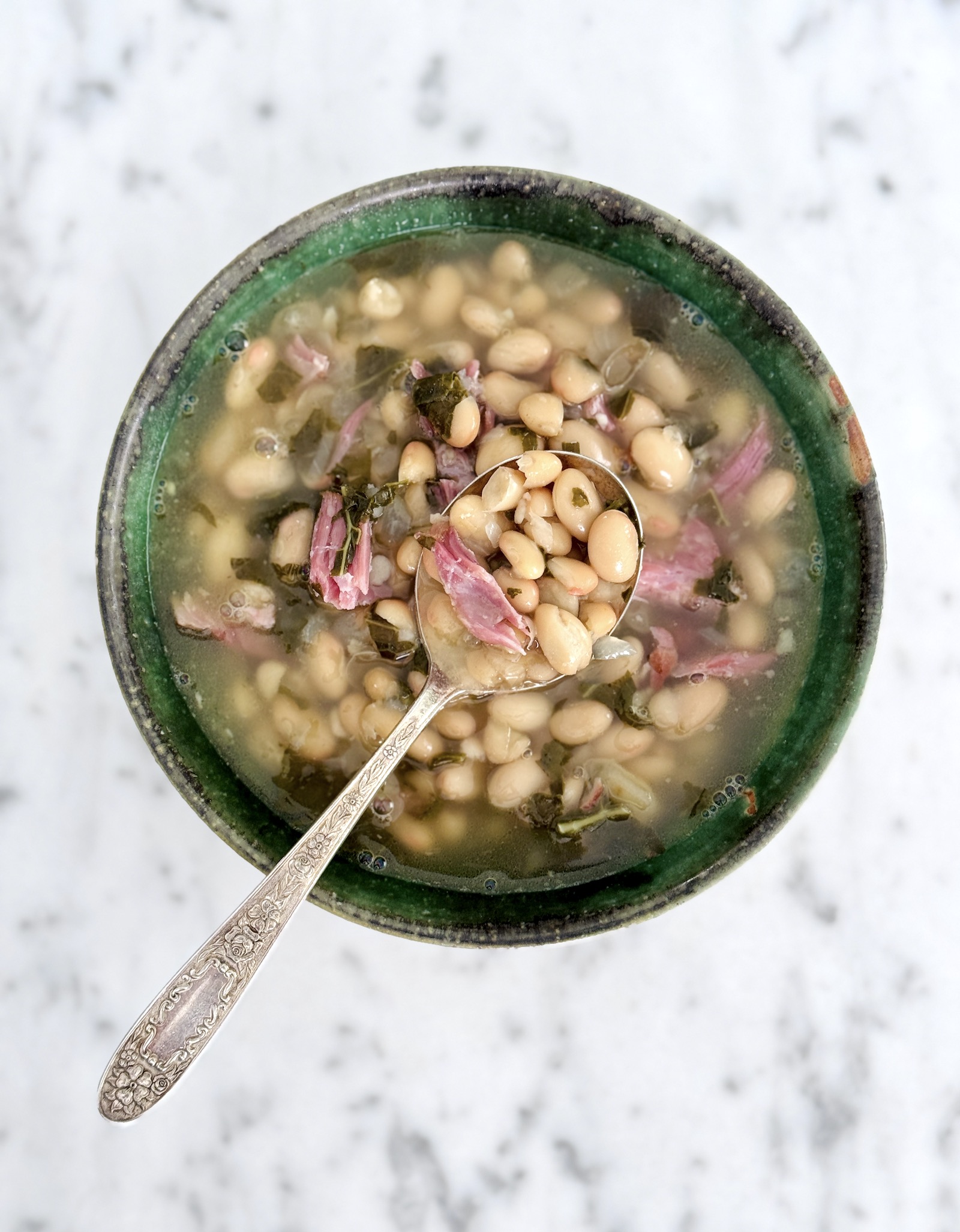 Bean and ham soup on a spoon above a green pottery bowl.